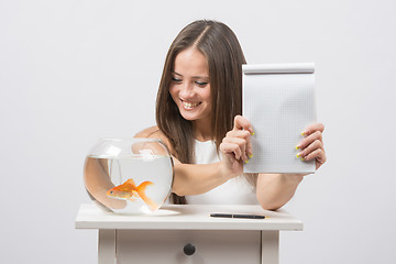 Image showing Girl shows a notebook with a record, standing next to an aquarium with goldfish