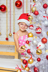 Image showing The girl in round glasses and a red Christmas hat hangs balls on a snowy New Years Christmas tree