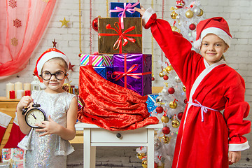 Image showing A girl holding a clock, a girl dressed as Santa Claus is standing at the bag with gifts