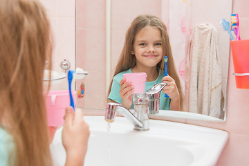 Image showing Happy little girl standing with toothbrush and cup and looks in the mirror