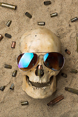 Image showing skull lying in the sand, scattered rifle and pistol cartridges. concept of war