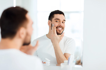 Image showing happy young man applying cream to face at bathroom