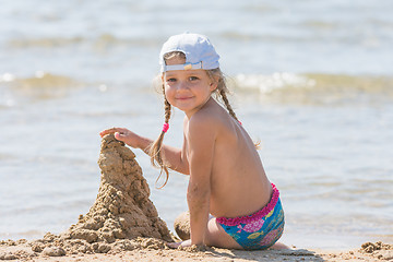 Image showing Five-year girl in a cap sits on the sand on the beach and building sand castle
