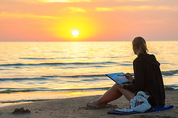 Image showing Young girl sitting on the beach, drawing on a sheet of watercolor sunset and looked at the setting sun