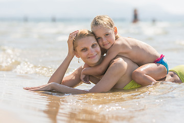 Image showing Mom and sits on her back the baby lying in the water on the sandy beach and happily look into the frame