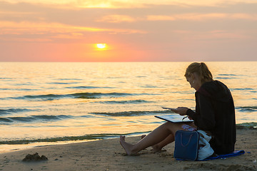 Image showing Young girl sitting on the beach and draw on a sheet of watercolor sunset