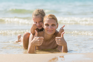 Image showing Mum with a daughter on his back lying on the beach and show. Girl and woman show thumbs up