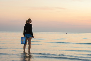 Image showing Young girl standing in the water on the beach with a landscape painted on a sheet of paper and brush