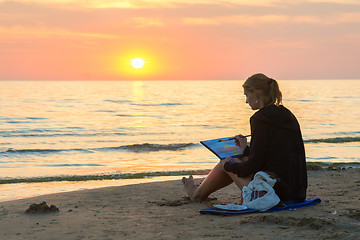 Image showing  Young girl sitting on the beach and draw on a sheet of watercolor sunset