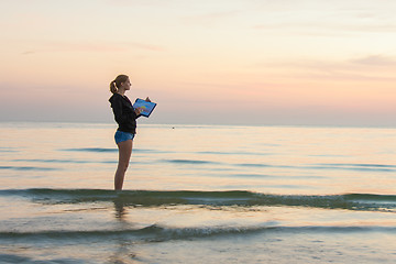 Image showing Young girl standing in the water on the beach, and draws on the colors of the sunset on a piece of paper