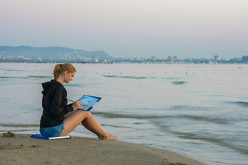 Image showing The young artist is sitting on the beach and drawing landscape with sunset