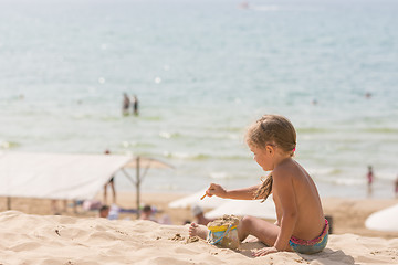 Image showing The little five year old girl sitting on the beach and playing with sand and toys on the background of the sea