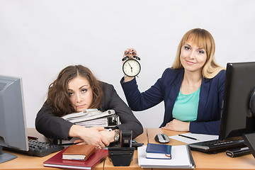 Image showing The girl in the office with a smile shows the time on the clock next to the exhausted colleague