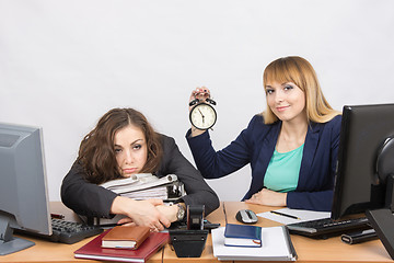 Image showing Two girls in the office at the end of the day, one with a smile, holding a clock, another weary lies on folders