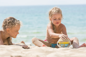 Image showing Two girls on the beach on a sunny day, playing with sand and a bucket on a background of the sea