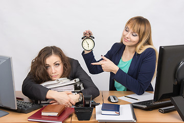 Image showing The girl in the office with a smile, holding a clock and looking at the tormented colleague