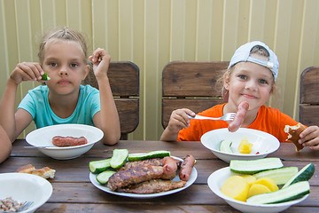 Image showing Two little girls at a wooden table in nature eating grilled sausages