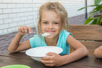 Image showing Satisfied six year old girl at the table in the courtyard eating porridge