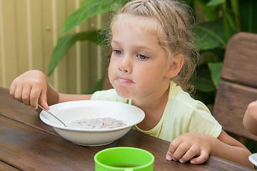 Image showing A girl of four years at a table outdoors thoughtfully eating porridge