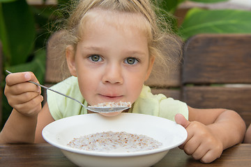 Image showing Four-year-girl at the table in the fresh air blowing on a spoonful of porridge