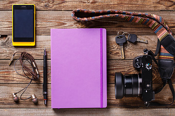 Image showing Overhead view of travel gear placed on wooden table. Mobile phone, earplugs, violet sketchbook, pencil, camera and purse. Flat lay top view.