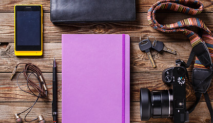 Image showing Overhead view of travel gear placed on wooden table. Mobile phone, earplugs, violet sketchbook, pencil, camera and purse. Flat lay top view.