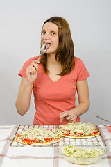 Image showing Girl sitting at a table in front of the two has not yet baked pizzas and licks the spoon