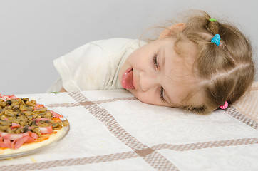 Image showing Little girl with protruding tongue rested her head on the table and looks at the pizza