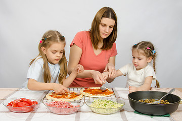 Image showing Mom helps younger daughter spread ketchup on a pizza, the eldest daughter, she is preparing a second pizza