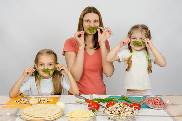 Image showing Young housewife with two daughters having fun holding sprig of parsley as a mustache at the kitchen table when sharing cooking
