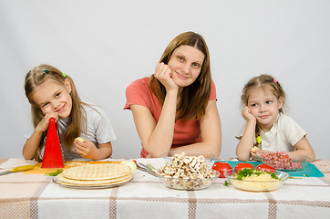 Image showing Satisfied mother with two daughters sitting resting his head in his hands at the table with the products for pizza