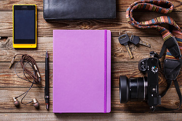 Image showing Overhead view of travel gear placed on wooden table. Mobile phone, earplugs, violet sketchbook, pencil, camera and purse. Flat lay top view.