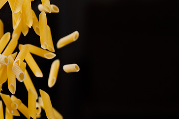 Image showing Falling penne pasta. Flying yellow raw macaroni over black background.