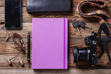 Image showing Overhead view of travel gear placed on wooden table. Mobile phone, earplugs, violet sketchbook, pencil, camera and purse. Flat lay top view.