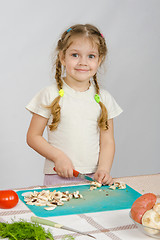 Image showing Little girl stands at the kitchen table, with a smile, looking into the frame and slice the mushrooms with a knife