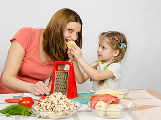 Image showing Six-year girl with pigtails giving mom a bite out of a piece of cheese at the kitchen table where they cook together