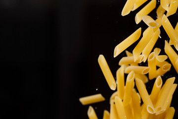 Image showing Falling penne pasta. Flying yellow raw macaroni over black background.