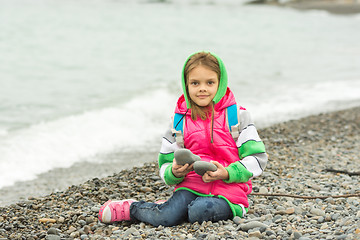 Image showing Seven-year girl sitting on a pebble beach in the warm clothes and looks in the frame