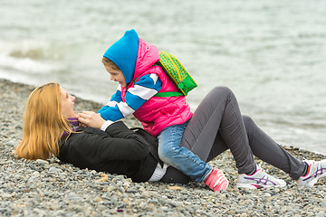 Image showing Five-year girl in warm clothes sits astride the mother who is lying on the pebble beach and sea fun looking at each other