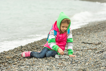 Image showing Seven-year girl sitting on a pebble beach in the warm clothing and with enthusiasm plays with stones