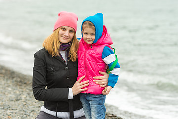 Image showing Mom and daughter in warm clothes hugging each other on the beach in cold weather and with a smile look in the frame