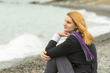 Image showing A girl sits on a pebble beach by the sea on a cloudy day in cold weather, and smiles enigmatically