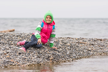 Image showing Warmly dressed smiling girl sitting on the pebble near the sea and looking at the frame