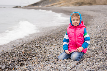 Image showing Five-year girl sits on a pebble beach in the warm bright clothes on a cloudy day with a smile looks in the frame