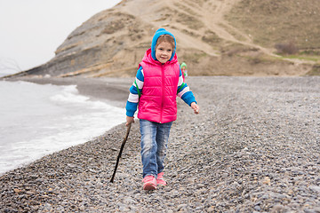 Image showing Five-year girl walking on the pebble beach in the warm bright clothes with a stick in his hand