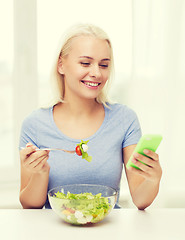 Image showing smiling woman with smartphone eating salad at home