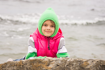 Image showing Warmly dressed girl looks out from behind a rock against the backdrop of the sea on a cold cloudy day