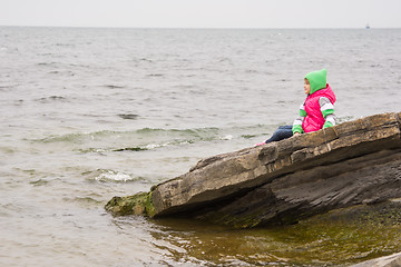 Image showing Warmly dressed little girl sitting on a piece of coastal cliffs and looking at the sea