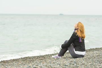 Image showing Young girl sitting on a pebble beach by the sea face to the sea breeze on a cloudy day