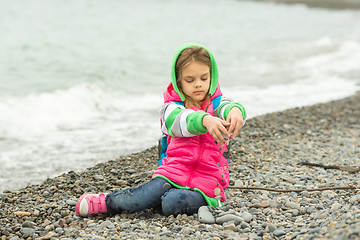 Image showing Seven-year girl sitting on a pebble beach in the warm clothing and pours out through her fingers small stones